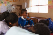 Seitenansicht eines jungen afrikanischen Schülers, der an seinem Schreibtisch sitzt, sich während eines Unterrichts in einer Township-Grundschule lehnt und zuhört, um ihn herum sitzen Mitschüler an ihren Schreibtischen und schreiben. — Stockfoto