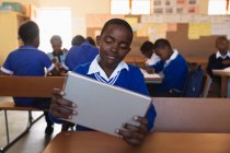 Vista frontal de cerca de un joven colegial africano sentado en su escritorio mirando una tableta y sonriendo durante una lección en un aula de la escuela primaria del municipio, en el fondo compañeros de clase están sentados en sus escritorios trabajando - foto de stock