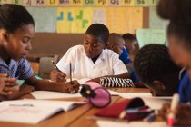 Vista frontal de un joven colegial africano sentado en su escritorio escribiendo durante una lección en un aula de la escuela primaria del municipio, a su alrededor compañeros de clase también están sentados en sus escritorios de escritura - foto de stock