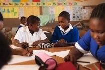 Vista frontal de dois jovens estudantes africanos sentados em uma mesa escrevendo e conversando durante uma aula em uma sala de aula da escola primária da cidade, em torno deles os colegas de classe também estão sentados em mesas escrevendo — Fotografia de Stock