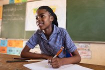 Вид спереди на молодую африканскую школьницу, сидящую за столом, улыбающуюся, пишущую в записной книжке и внимательно слушающую во время урока в городском классе начальной школы — стоковое фото
