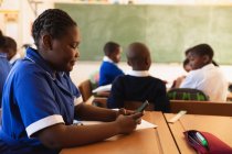Vista lateral de cerca de una joven colegiala africana sentada en su escritorio usando un teléfono inteligente y sonriendo en un aula en una escuela primaria del municipio . - foto de stock