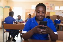 Взгляд крупным планом на молодую африканскую школьницу, сидящую за партой со смартфоном и улыбающуюся в классе местной школы . — стоковое фото