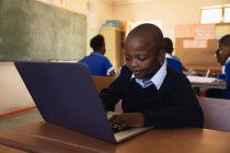 Vista frontale da vicino di un giovane scolaro africano seduto alla sua scrivania con un computer portatile e sorridente durante una lezione in una classe della scuola elementare cittadina, sullo sfondo i compagni di classe sono seduti alle loro scrivanie a lavorare — Foto stock