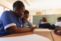 Vista lateral de perto de duas jovens alunas africanas sentadas em uma mesa usando um smartphone juntas e sorrindo em uma sala de aula em uma escola primária da cidade . — Fotografia de Stock