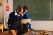 Seitenansicht von zwei jungen afrikanischen Schülern, die während einer Unterrichtspause in einer Township-Grundschule auf Stühlen sitzen und auf einen Tablet-Computer schauen — Stockfoto