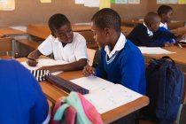 Vista frontal close-up de dois jovens estudantes africanos sentados em uma mesa escrevendo e conversando durante uma aula em uma sala de aula da escola primária da cidade, nos colegas de escola de fundo também estão sentados em mesas escrevendo — Fotografia de Stock
