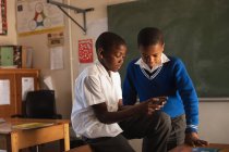 Vista lateral de cerca de dos jóvenes escolares africanos mirando un teléfono inteligente durante un descanso de las lecciones en un aula de la escuela primaria del municipio - foto de stock