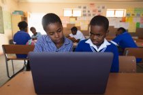 Vue de face gros plan d'un jeune écolier africain et d'une écolière assis à un bureau à l'aide d'un ordinateur portable ensemble pendant une leçon dans une classe de l'école élémentaire d'un canton, en arrière-plan, des camarades de classe sont assis à leur bureau travaillant — Photo de stock