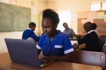 Vista frontal de cerca de una joven colegiala africana sentada en un escritorio usando una computadora portátil y sonriendo durante una lección en un aula de la escuela primaria del municipio, en el fondo los compañeros de clase están sentados en sus escritorios trabajando - foto de stock