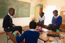 Підвищений вид з чотирьох молодих африканських школярів сидять навколо столу говорити під час перерви з уроків в містечку початкової школи школі, три з них сидять на спинах стільців — стокове фото