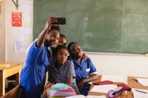 Vista frontal de cerca de un grupo de jóvenes colegialas africanas divirtiéndose posando y tomando selfies con un teléfono inteligente durante un descanso de las lecciones en un aula de la escuela primaria del municipio - foto de stock