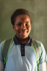 Retrato de cerca de una joven colegiala africana vestida con su uniforme escolar y su mochila, mirando directamente a la cámara sonriendo, en una escuela primaria del municipio - foto de stock