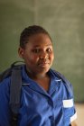 Ritratto da vicino di una giovane studentessa africana che indossa l'uniforme scolastica e lo zaino, guardando dritto verso la telecamera sorridente, in una scuola elementare cittadina — Foto stock
