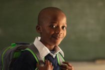 Retrato de cerca de un joven colegial africano vestido con su uniforme escolar y su mochila, mirando directamente a la cámara sonriendo, en una escuela primaria del municipio - foto de stock