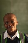 Ritratto da vicino di un giovane scolaro africano che indossa l'uniforme scolastica e lo zaino, guardando dritto davanti alla telecamera sorridente, in una scuola elementare cittadina — Foto stock