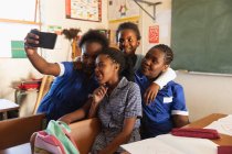 Vista frontal de cerca de un grupo de jóvenes colegialas africanas divirtiéndose posando y tomando selfies con un teléfono inteligente durante un descanso de las lecciones en un aula de la escuela primaria del municipio - foto de stock