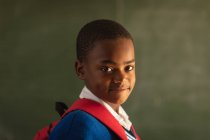 Retrato de cerca de un joven colegial africano vestido con su uniforme escolar y su mochila, mirando directamente a la cámara sonriendo, en una escuela primaria del municipio - foto de stock