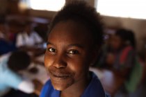 Erhabenes Porträt eines jungen afrikanischen Mädchens, das lächelnd direkt in die Kamera blickt, in einer Township-Grundschule, während ihre Klassenkameraden an einem Schreibtisch im Hintergrund sitzen — Stockfoto