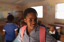Portrait close up of a young African schoolgirl wearing her school uniform and schoolbag, looking straight to camera smiling, at a township elementary school with classmates in the background — Stock Photo