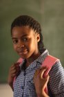 Retrato de cerca de una joven colegiala africana vestida con su uniforme escolar y su mochila, mirando directamente a la cámara sonriendo, en una escuela primaria del municipio - foto de stock
