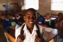 Retrato de cerca de un joven colegial africano vestido con su uniforme escolar y su mochila, mirando directamente a la cámara sonriendo, en una escuela primaria del municipio con compañeros de clase sentados en escritorios en el fondo - foto de stock