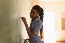 Vue de côté gros plan d'une jeune écolière africaine debout à l'avant de la classe écrivant sur le tableau noir lors d'une leçon dans une classe de l'école élémentaire d'un canton — Photo de stock
