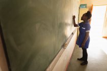 Vista lateral de una joven colegiala africana de pie al frente de la clase escribiendo en la pizarra durante una lección en un aula de la escuela primaria del municipio - foto de stock