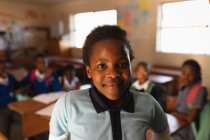 Retrato de cerca de una joven colegiala africana vestida con su uniforme escolar y su mochila, mirando directamente a la cámara sonriendo, en una escuela primaria del municipio con compañeros de clase sentados en escritorios en el fondo - foto de stock