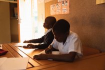 Vista lateral de cerca de dos jóvenes escolares africanos sentados en un escritorio trabajando durante una lección en una escuela primaria del municipio - foto de stock