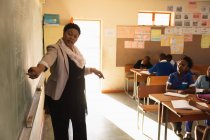 Front view close up of a middle aged African female school teacher standing at the front of the class pointing at the blackboard with pupils watching from their desks during a lesson in a township elementary school classroom — Stock Photo