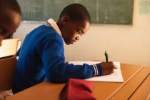 Nahaufnahme eines jungen afrikanischen Schülers, der an einem Schreibtisch sitzt und während eines Unterrichts in einer Township-Grundschule schreibt — Stockfoto