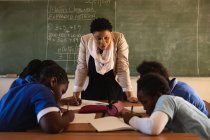 Front view close up of a middle aged African female school teacher standing at the front of the class in front of the blackboard and leaning forward to watch her pupils writing at their desks during a lesson in a township elementary school classroom — Stock Photo