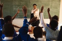 Вид спереди африканской учительницы средних лет, стоящей перед классом перед доской, с видом сзади учеников, сидящих за партами с поднятыми вверх руками во время урока — стоковое фото
