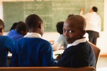 Side view close up of a young African schoolboy sitting at his desk and turn around, looking to camera and smiling during a lesson in a township elementary school classroom . — стоковое фото
