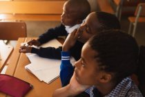 Vista lateral elevada de cerca de una joven colegiala africana y dos escolares sentados en un escritorio escuchando durante una lección en un aula de la escuela primaria del municipio - foto de stock