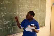 Front view close up of a young African schoolgirl standing at the front of the class holding a book and writing on the blackboard during a lesson in a township elementary school classroom — Stock Photo