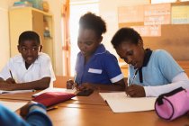 Frontansicht eines jungen afrikanischen Schülers und zweier Schulmädchen, die an Schreibtischen sitzen und während einer Unterrichtsstunde in einem Klassenzimmer der Township-Grundschule schreiben — Stockfoto