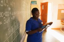 Vista laterale da vicino di una giovane studentessa africana in piedi davanti alla lavagna che tiene un libro durante una lezione in una classe della scuola elementare cittadina — Foto stock