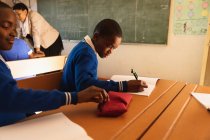 Vue latérale de deux jeunes écoliers africains assis à un bureau travaillant pendant une leçon dans une classe de l'école élémentaire d'un canton, en arrière-plan l'enseignant aide certains camarades de classe à leur bureau — Photo de stock
