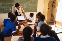 Вид спереди на учителя африканской школы средних лет, стоящего перед доской и показывающего своим ученикам ноутбук во время урока в классе начальной школы.
. — стоковое фото