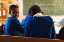 Seitenansicht von einem jungen afrikanischen Schüler, der an seinem Schreibtisch sitzt und sich umdreht, in die Kamera schaut und während einer Unterrichtsstunde lächelt. — Stockfoto