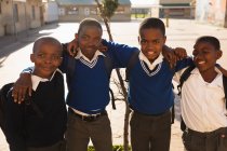 Портрет с четырьмя молодыми африканскими школьниками, стоящими с обнимающимися руками и улыбающимися перед камерой в школьном дворе начальной школы. — стоковое фото