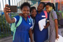 Vista frontal de cerca de un grupo de jóvenes colegialas africanas que se divierten posando y tomando selfies con un teléfono inteligente en un patio de recreo de la escuela primaria del municipio - foto de stock