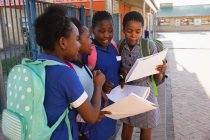 Vista lateral de cerca de cuatro jóvenes colegialas africanas que llevan bolsos escolares, mirando los libros escolares en el patio de recreo de una escuela primaria del municipio - foto de stock