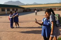 Vue latérale de deux jeunes écolières africaines regardant un smartphone ensemble dans une aire de jeux de l'école élémentaire du canton. En arrière-plan, deux de leurs camarades de classe regardent leurs livres scolaires — Photo de stock