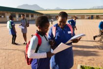 Vista lateral de cerca de dos jóvenes colegialas africanas que llevan bolsos escolares, mirando un libro escolar en el patio de recreo de una escuela primaria del municipio . - foto de stock