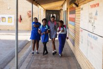 Vue de face de quatre jeunes écolières africaines portant des cartables souriants et marchant les bras liés dans l'aire de jeux d'une école élémentaire d'un canton — Photo de stock