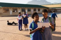 Vue de face gros plan de deux jeunes écolières africaines portant des cartables, regardant un livre d'école dans la cour de récréation d'une école élémentaire d'un canton . — Photo de stock