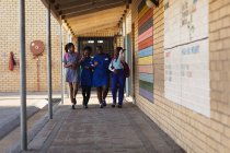 Вид спереди на четырех молодых африканских школьниц, одетых в школьные сумки, улыбающихся и идущих с руками, связанных на детской площадке начальной школы. — стоковое фото